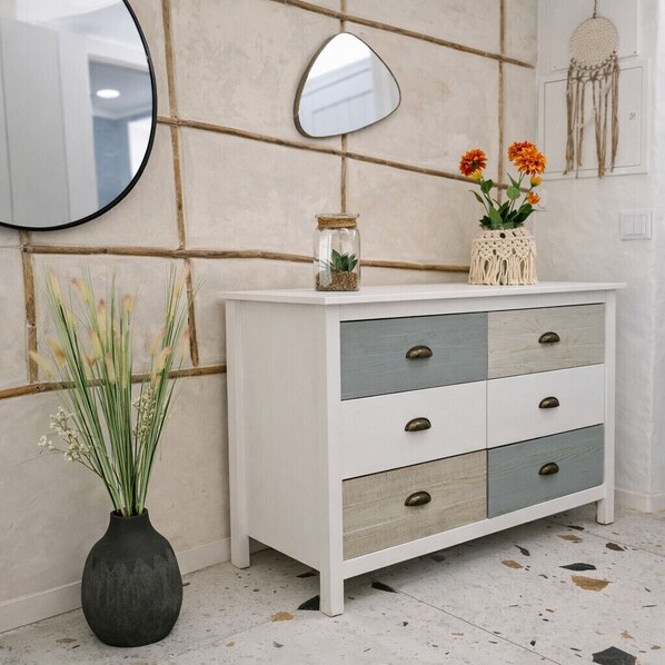 Une commode blanche avec des tiroirs, un miroir rond, un vase avec des fleurs et un attrape-rêves accroché au mur.