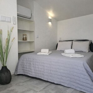 A modern bedroom with a grey bed, white pillows, a bedside table, a wall-mounted air conditioner, and a small shelf with decorative items.