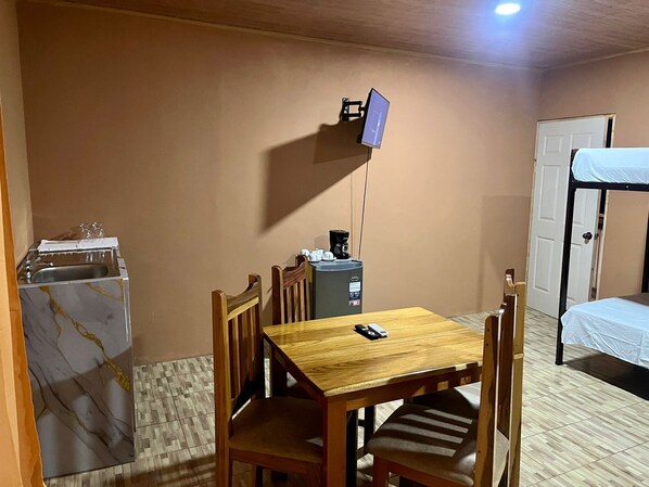 Une pièce compacte comprenant une table en bois, des chaises, une petite kitchenette avec un évier, une machine à café et un téléviseur fixé au mur.