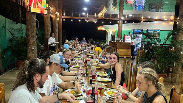 A group of people dining at a wooden table in an outdoor setting with string lights overhead. - Luisito steak house & hotel (La Fortuna)