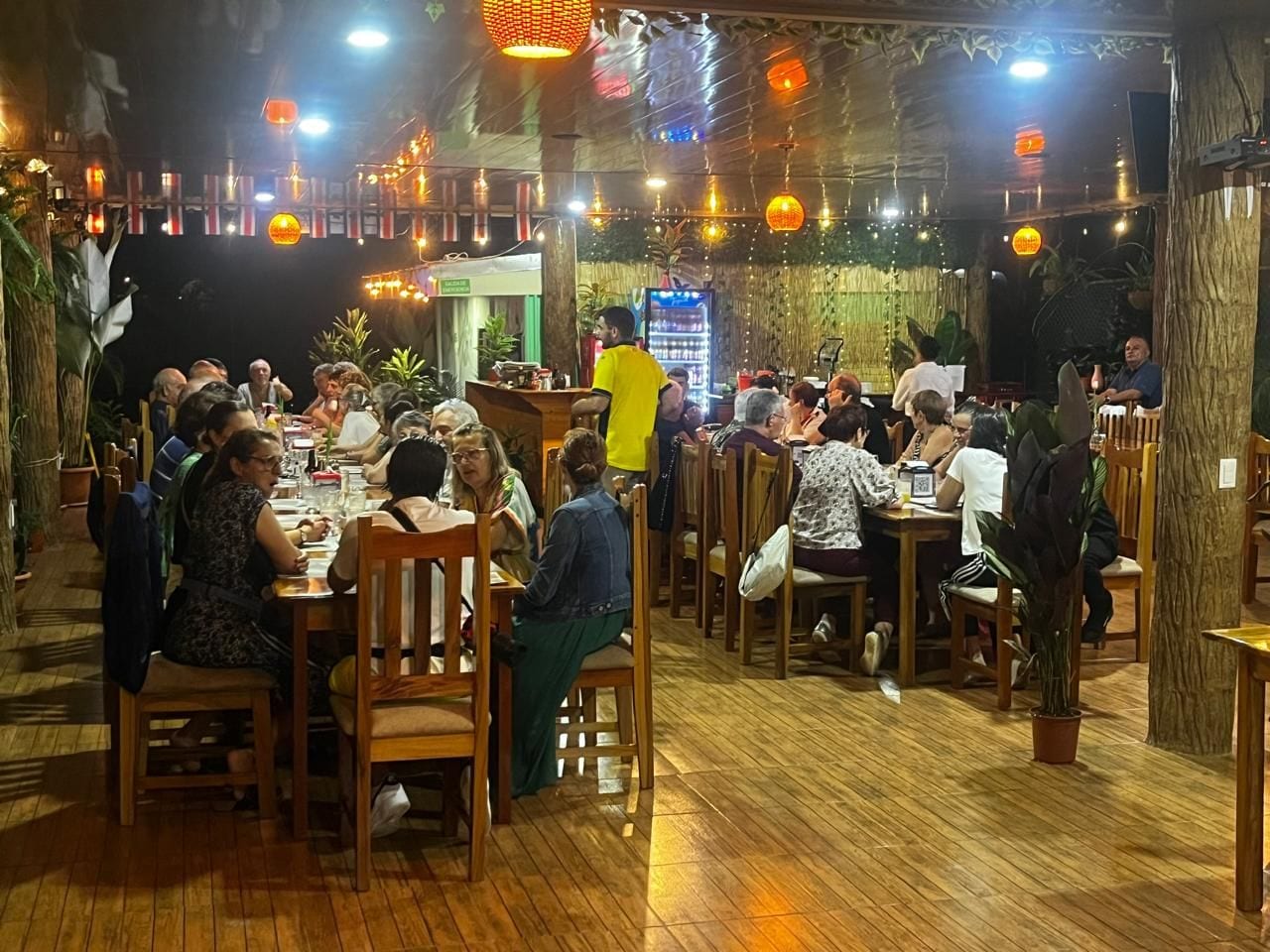 Un restaurante con mesas y sillas de madera, rodeado de macetas con plantas y faroles colgantes.