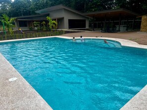 Una piscina con agua azul cristalina, rodeada por una terraza empedrada y un edificio con grandes ventanales.