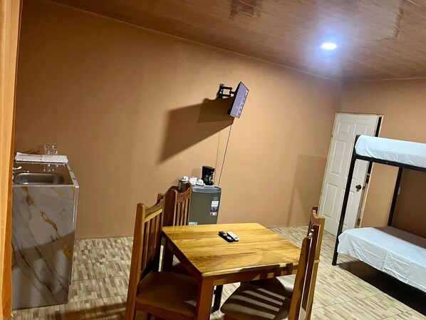 Una habitación compacta con una mesa de madera, una cama litera, una pequeña cocina y un televisor montado en la pared.