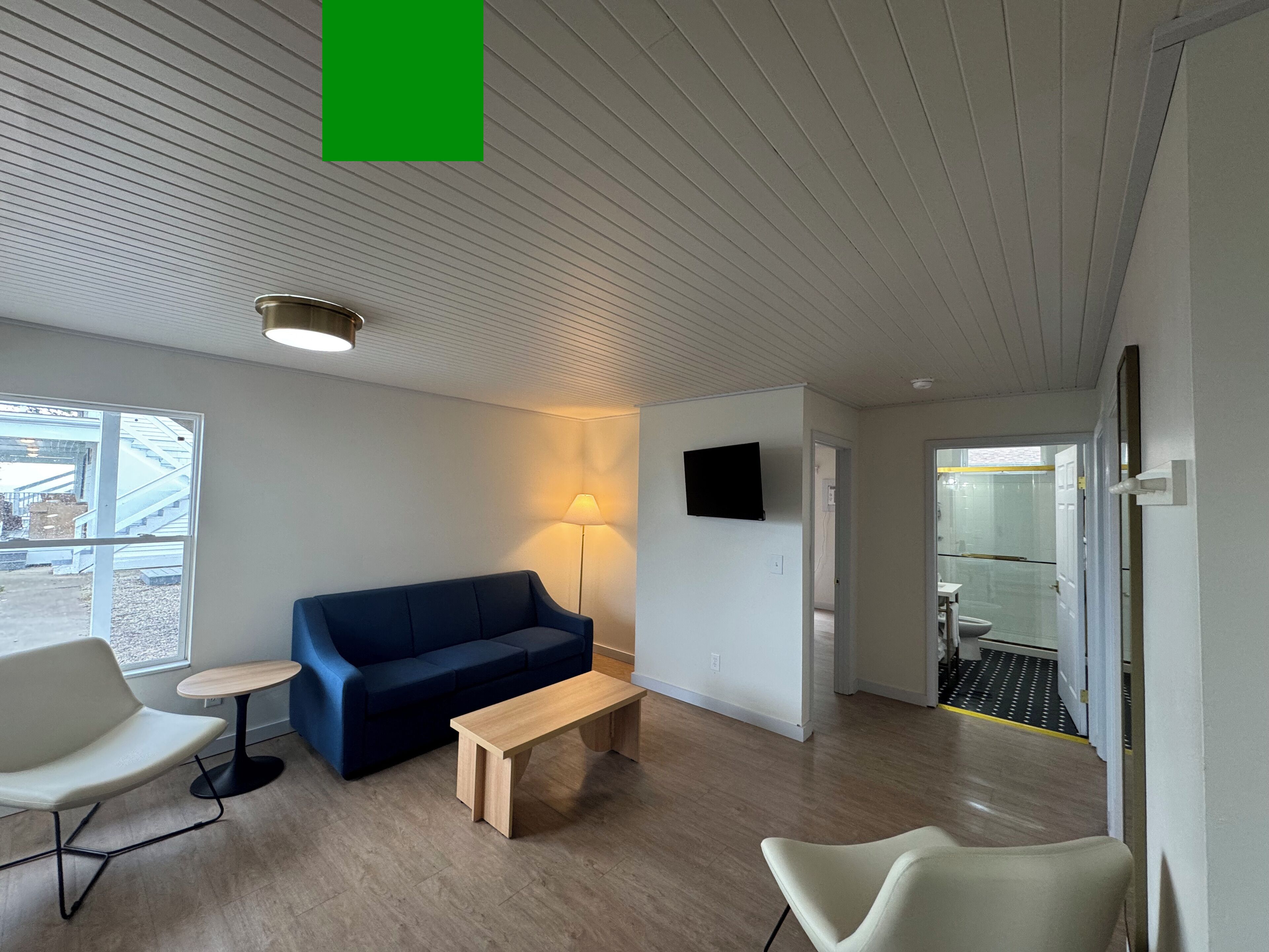 Un espace d’attente moderne avec un canapé bleu, un banc en bois et une chaise blanche. Une lampe est fixée au mur, et un téléviseur est installé au-dessus du canapé.