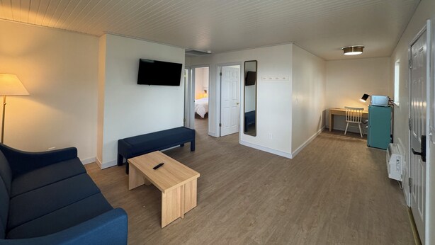 A modern hotel room with a blue sofa, a wooden coffee table, a flat-screen TV mounted on the wall, a mirror, and a kitchenette area with a microwave and a small fridge.