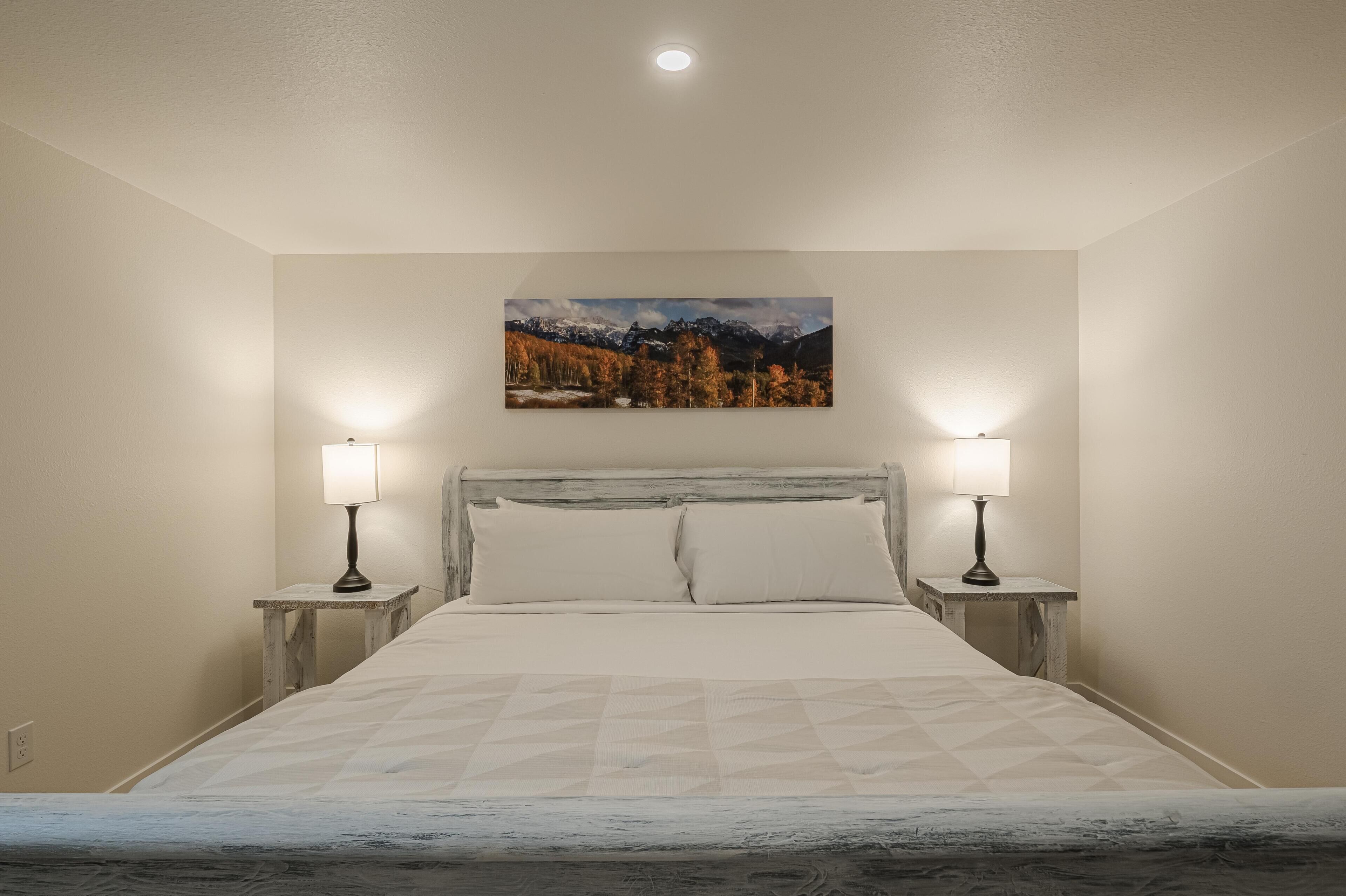 Ein Schlafzimmer mit einem Bett, zwei Nachttischen mit Lampen, einem Wandbild von einer Berglandschaft und einer Deckenleuchte.