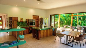 A modern kitchen with a central island, dining area with chairs, and a large window offering a view of greenery. - Charming country house in  Playa Órganos Paquera in the middle of nature (Paquera)