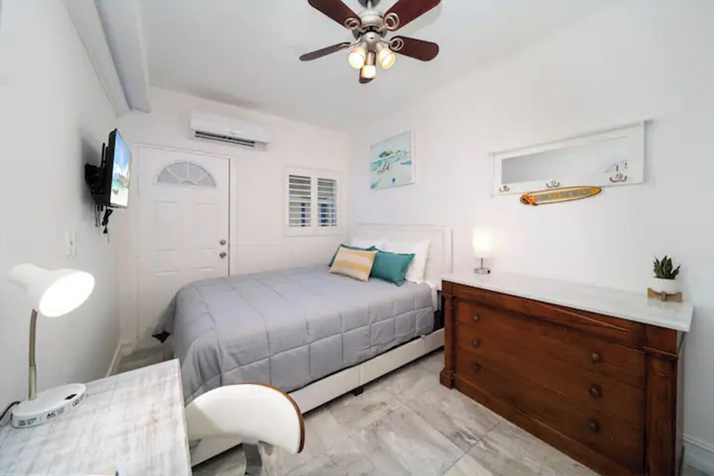 Een slaapkamer met een bed, nachtkastje, lamp, dressoir, plafondventilator en een wandairconditioner.