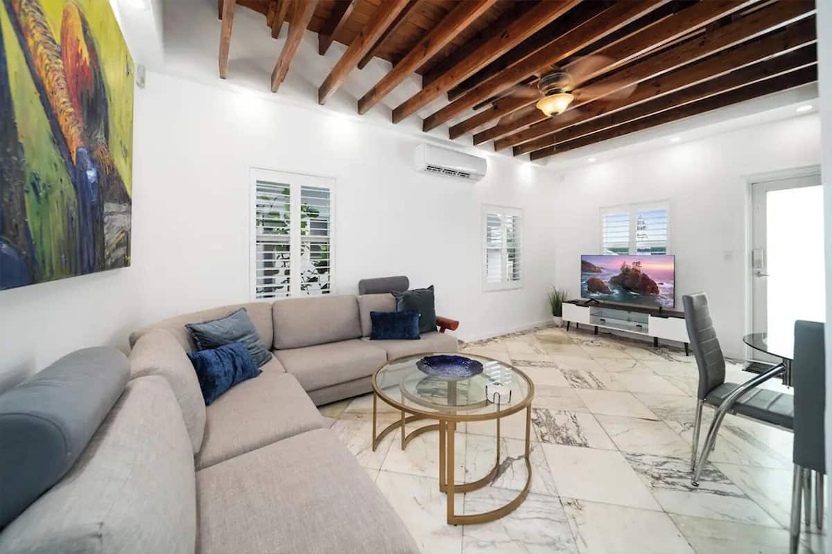 Una sala moderna con un sofá color beige, una mesa de centro de vidrio y un televisor sobre un mueble.