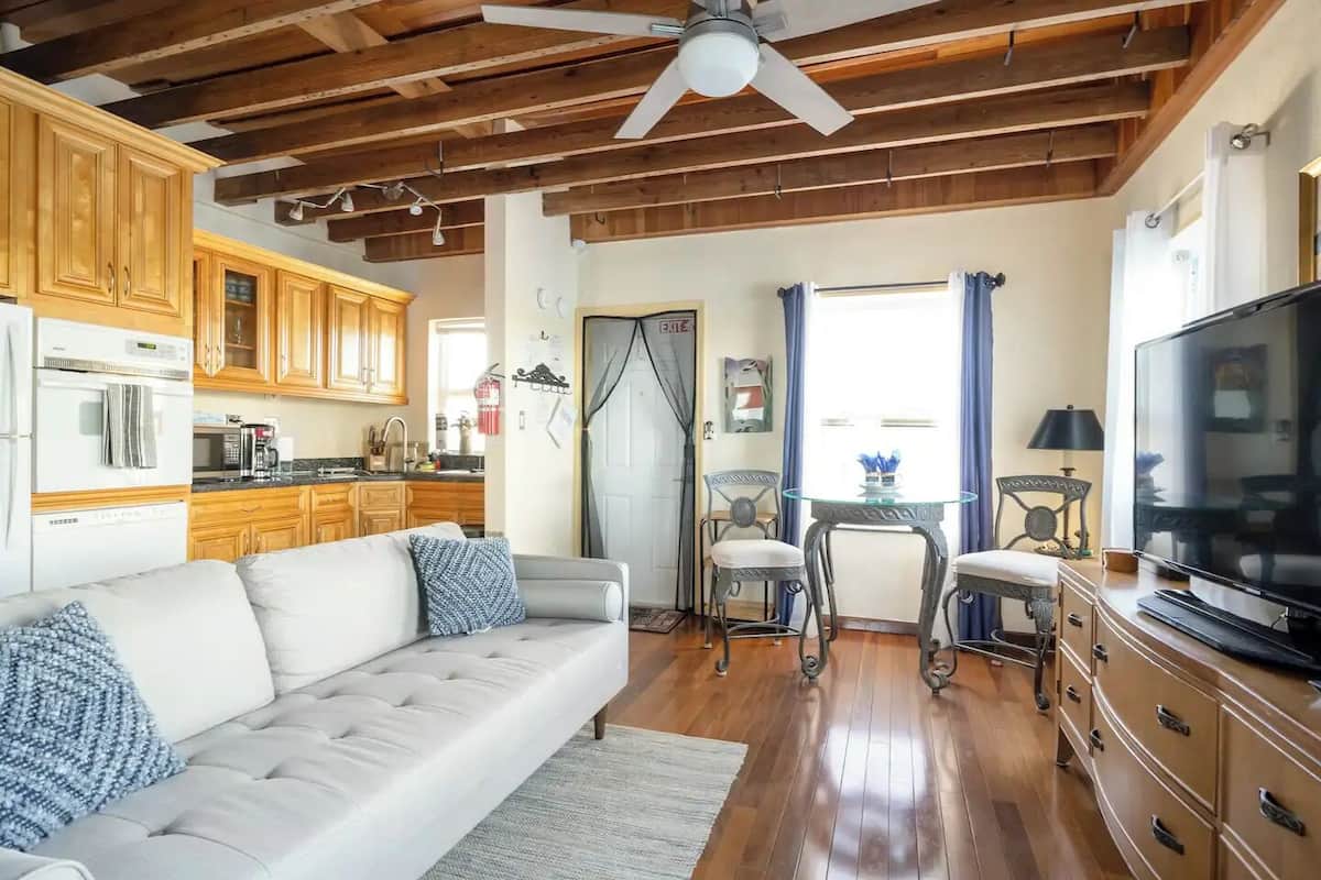 Una sala de estar con sofá, sillas, televisor, armarios de madera y una zona de cocina.