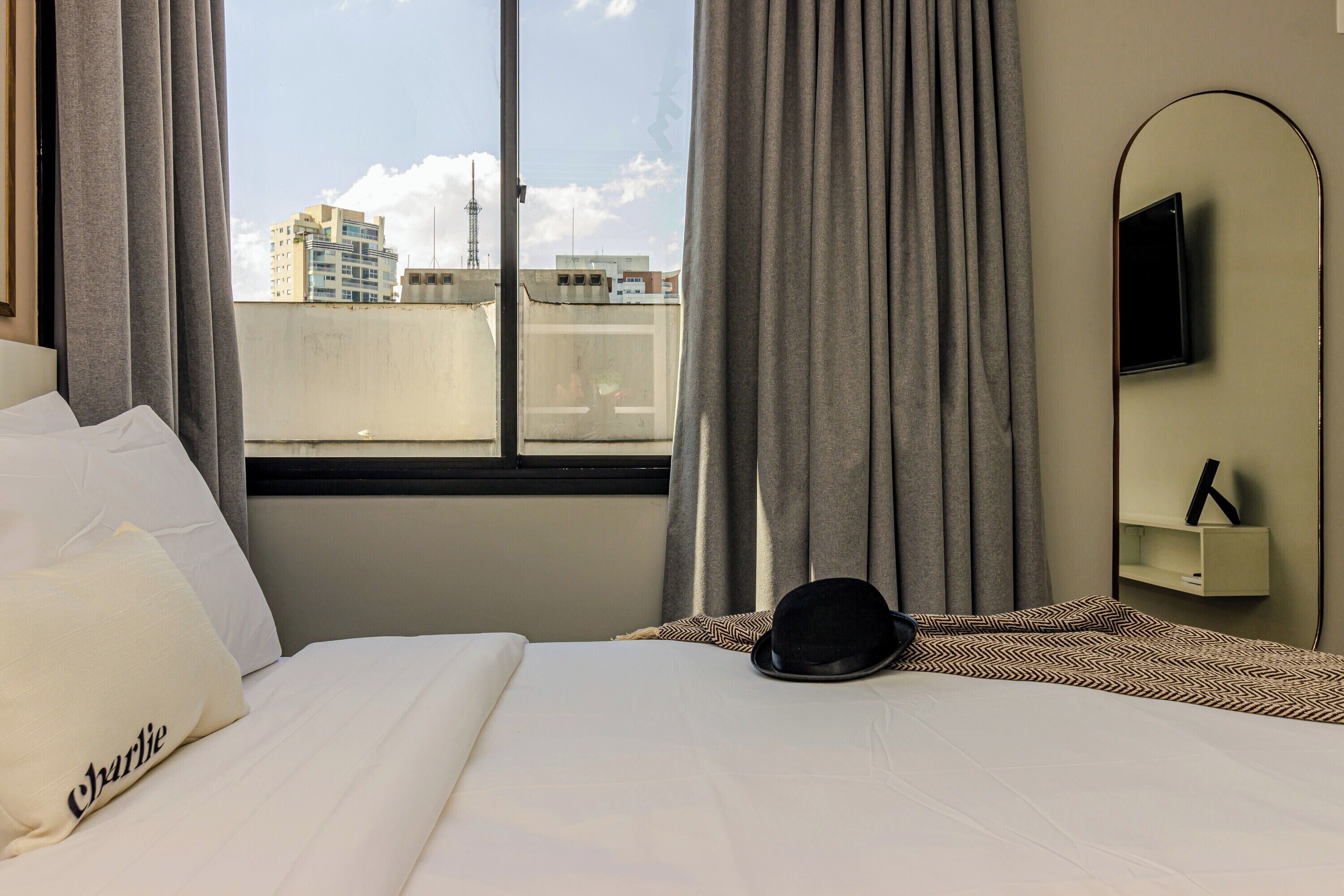 Quarto de hotel com uma cama, um chapéu preto, uma televisão na parede e vista para prédios pela janela.