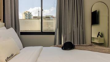 Quarto de hotel com uma cama, um chapéu preto, uma televisão na parede e vista para prédios pela janela.