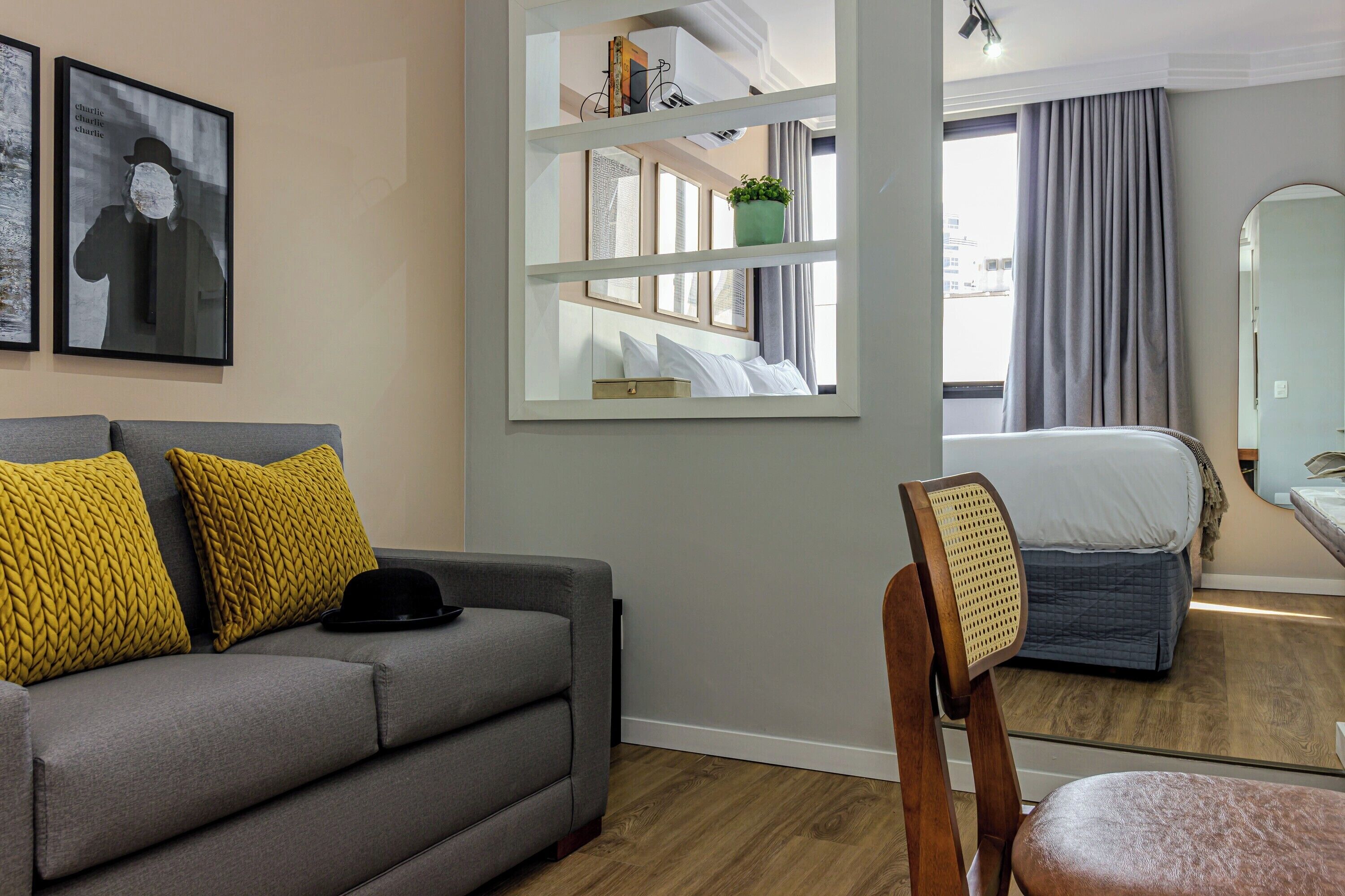 A hotel room with a grey sofa, yellow cushions, a wooden chair, a bed with a grey duvet, a mirror, and a window with curtains.