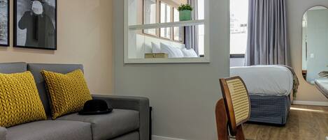 A hotel room with a grey sofa, yellow cushions, a wooden chair, a bed with a grey duvet, a mirror, and a window with curtains.