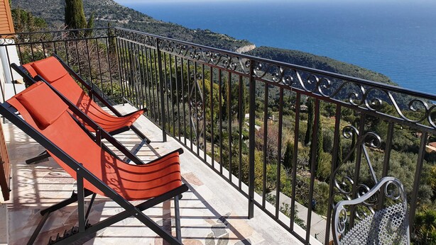 Un balcón con dos sillones naranjas y una silla de metal, con vistas a un paisaje costero que incluye mar y colinas.