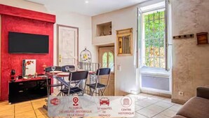A living room with a red accent wall, a flat-screen TV, a dining area with chairs, and a window with a view of greenery. - Le Capouane - The Tower of Augustus (La Turbie)