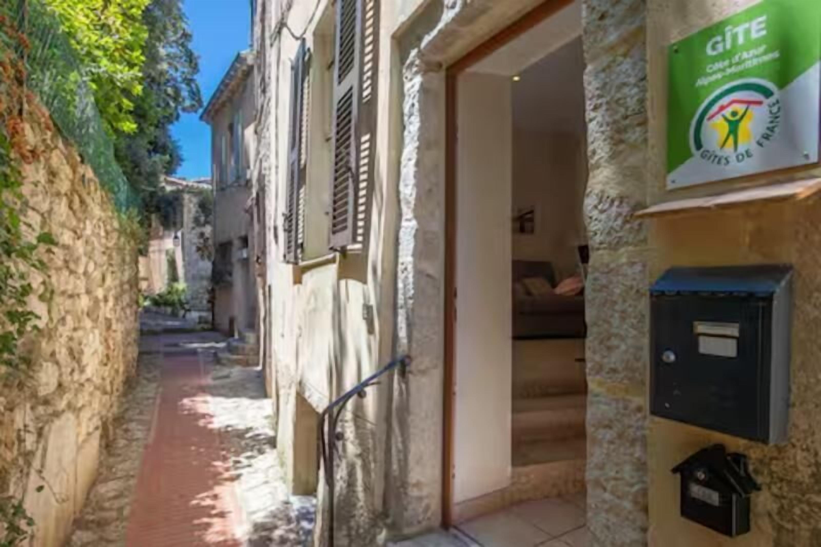 A narrow alleyway with stone walls and a red pathway. A sign for 'Gîte' is visible on the right, indicating a lodging option.