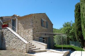 A stone building with a staircase leading to a door, a fenced area, and a tall tree.