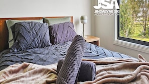 Un dormitorio con cama, almohadas, una colcha, una lámpara de noche y un ventanal con vista a follaje.