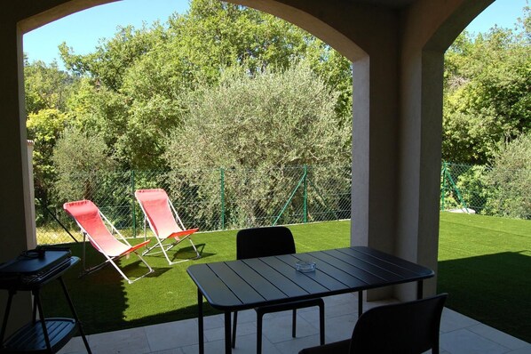 Une terrasse extérieure avec une table et des chaises, entourée d’une pelouse verdoyante et d’une haie.