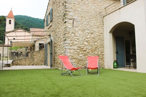 Un edificio de piedra con un patio cubierto de césped, dos sillas rojas y una torre de campanas al fondo.
