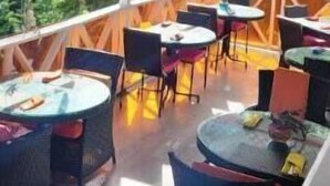 Un área de estar al aire libre con mesas y sillas redondas, un ventilador de techo y vista a la vegetación.