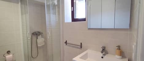 Un baño con ducha envuelta en vidrio, un lavabo blanco con dispensador de jabón de madera, y un espejo sobre el lavabo.