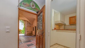 A hallway leading to a kitchen with a tiled floor, a wooden door with a decorative glass panel, and a view of a courtyard with a brick floor. - Le Du Guet - Augustus Tower (La Turbie)