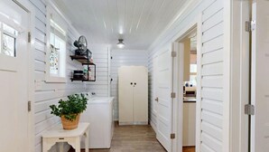 Een smalle gang met witte houten wanden en een houten vloer. Er staat een klein tafeltje met een plant in pot, een wasmachine, en een ventilator is aan de muur bevestigd. Aan het einde van de gang is een deur zichtbaar.