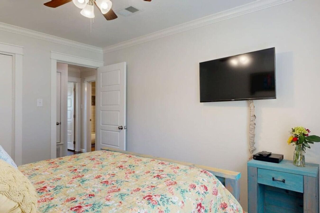 Un dormitorio con cama, televisión montada en la pared, una mesita con un florero y ventilador de techo.
