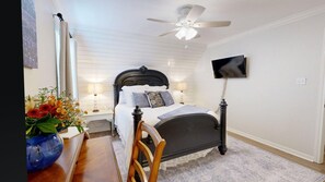 Un dormitorio con cama, mesitas de noche, televisor, ventilador de techo y ventana con cortinas.