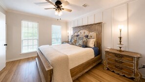 Une chambre à coucher avec un lit en bois, un ventilateur de plafond, une commode et deux fenêtres équipées de stores.