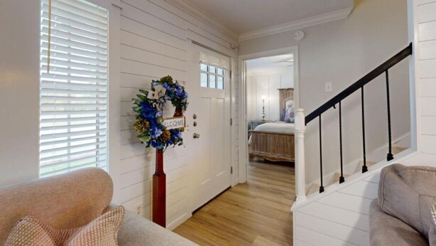 A hallway with a wooden floor, white walls, and a staircase with black railings. A welcome wreath is on the wall, and a bed is visible in the background.