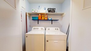 A laundry room with two front-loading washing machines, a shelf above with cleaning supplies, and a rack with bags. - Central Cottage Charm - Pet Friendly! Very Clean! (College Station)