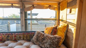 Interior acogedor de una cabaña con un banco acolchado, una ventana con cortinas y vistas a una terraza de madera.