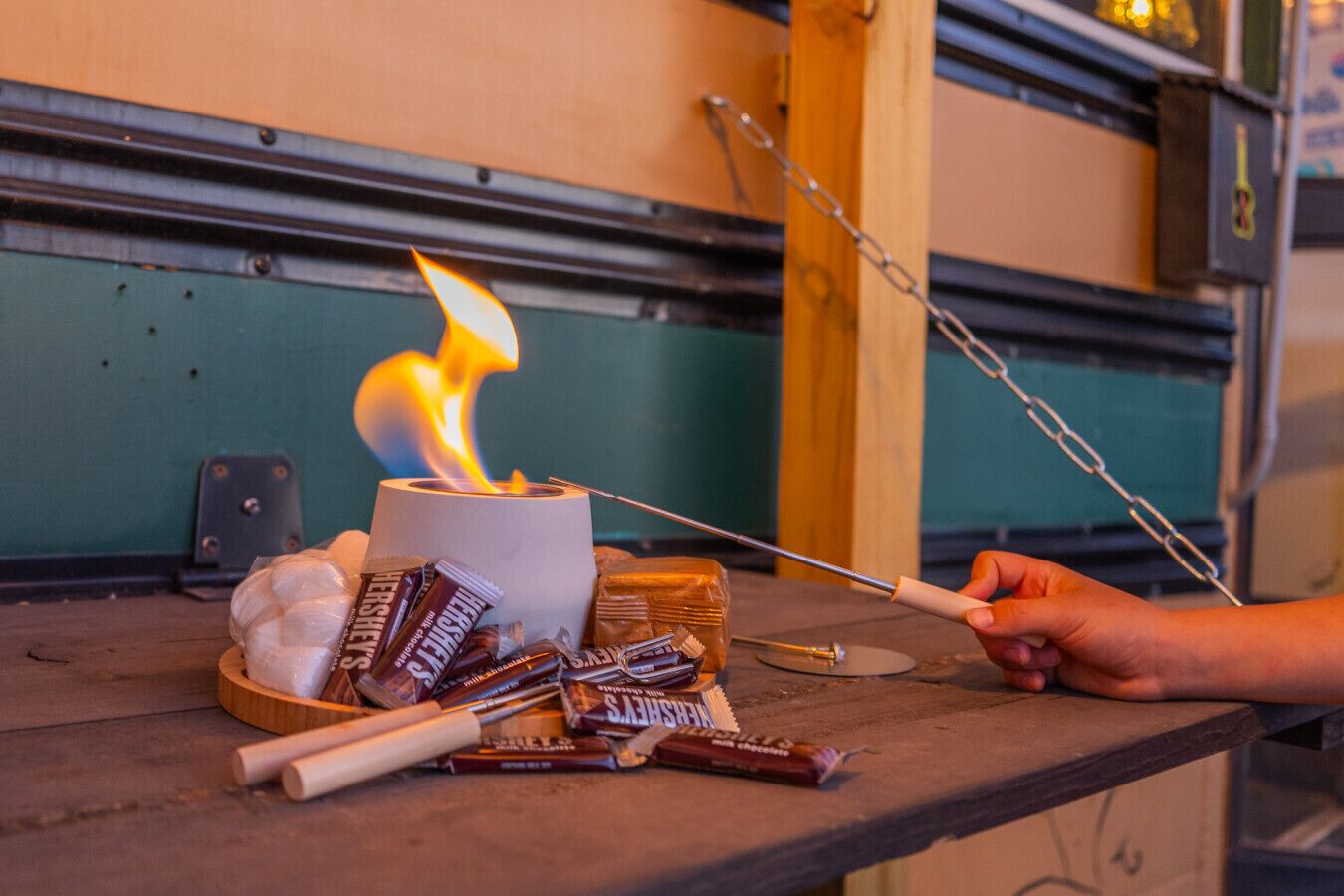 A person roasting marshmallows over a campfire with a stick, surrounded by chocolate bars and marshmallows on a wooden table.