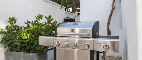 A stainless steel barbecue grill with four burners and a side burner, placed outdoors next to a concrete planter with green foliage.