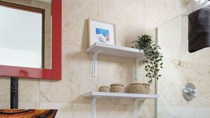 Un baño con inodoro blanco, tocador rojo con cajones, cabina de ducha de vidrio y estantes con cestas y una planta.