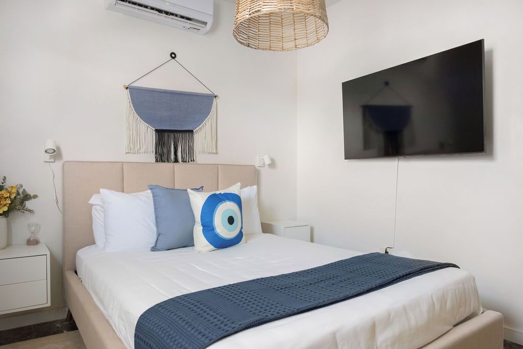 Ein ordentlich eingerichtetes Schlafzimmer mit Bett, Nachttischen, einem an der Wand montierten Fernseher und einer Wanddeko aus Makramee.