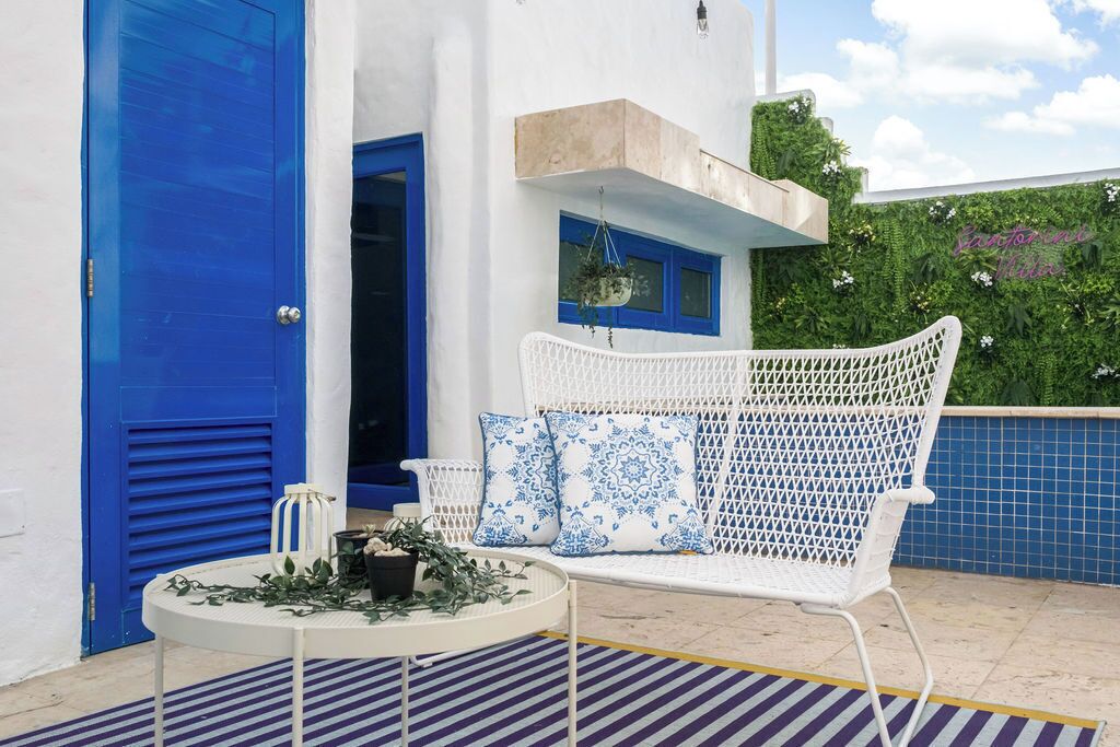 A white outdoor seating area with blue and white patterned cushions, a round table with a plant, and a blue door.