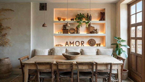 Un coin repas avec une table et des chaises en bois, un luminaire suspendu et des étagères ornées d’objets décoratifs et du mot « AMOR ».