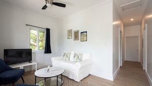 A living room with a white sofa, a TV on a stand, a ceiling fan, and a window with curtains. - Laughing Bird Suites (Governor's Harbour)