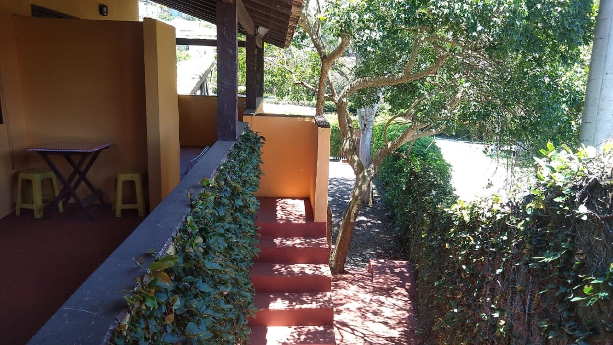 A covered outdoor area with a staircase leading to a higher level, surrounded by greenery and a clear view of the street.