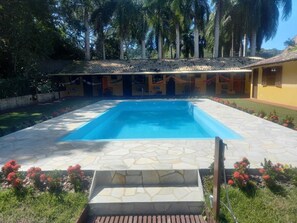 A swimming pool with stone tiles and a wooden deck in front, surrounded by a garden with red flowers and palm trees. - A Casa do Pablo (Buzios)