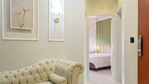 Ein Flur mit einem gesteppten Sofa, gerahmten Blumenbildern, einer Wandlampe und einer Tür zu einem Schlafzimmer mit Bett und Nachttisch.