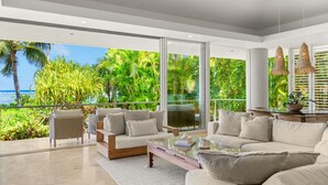 Una sala de estar moderna con un gran ventanal que da a un balcón y vistas al océano y a la vegetación exuberante.