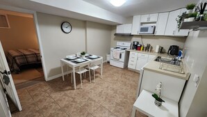 Une cuisine compacte avec des meubles blancs, un four à micro-ondes, une cuisinière et un évier. On y trouve une table à manger avec des chaises et une horloge au mur.
