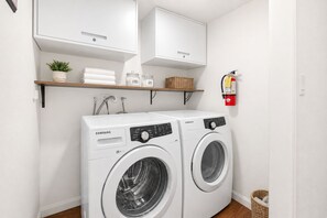 Ein Waschraum mit zwei Samsung Waschmaschinen (Frontlader), einem Feuerlöscher und Regalen mit Handtüchern und Körben.