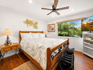 Une chambre avec un lit en bois, un ventilateur de plafond, une fenêtre donnant sur des arbres, une lampe fixée au mur et une étagère remplie de livres.
