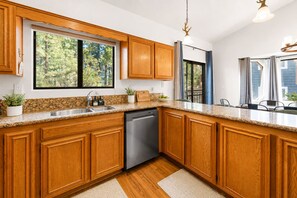 Une cuisine dotée de meubles en bois, d’un lave-vaisselle, de plans de travail en granit et d’une fenêtre donnant sur la forêt.